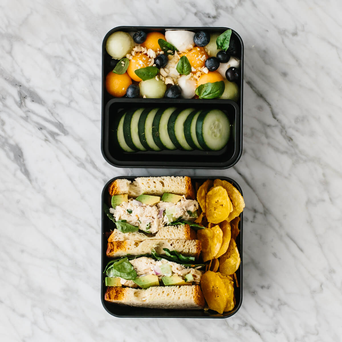 Bento box filled with tuna salad sandwich, fruit salad and chips.