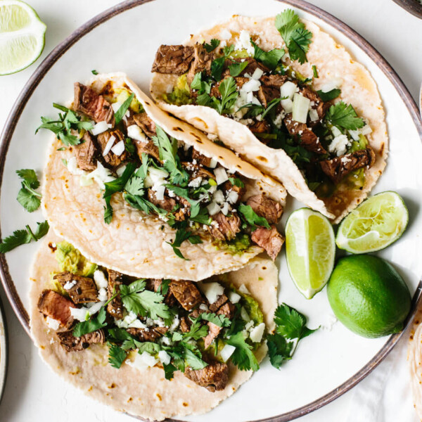 A plate of carne asada tacos next to tortillas and cheese.