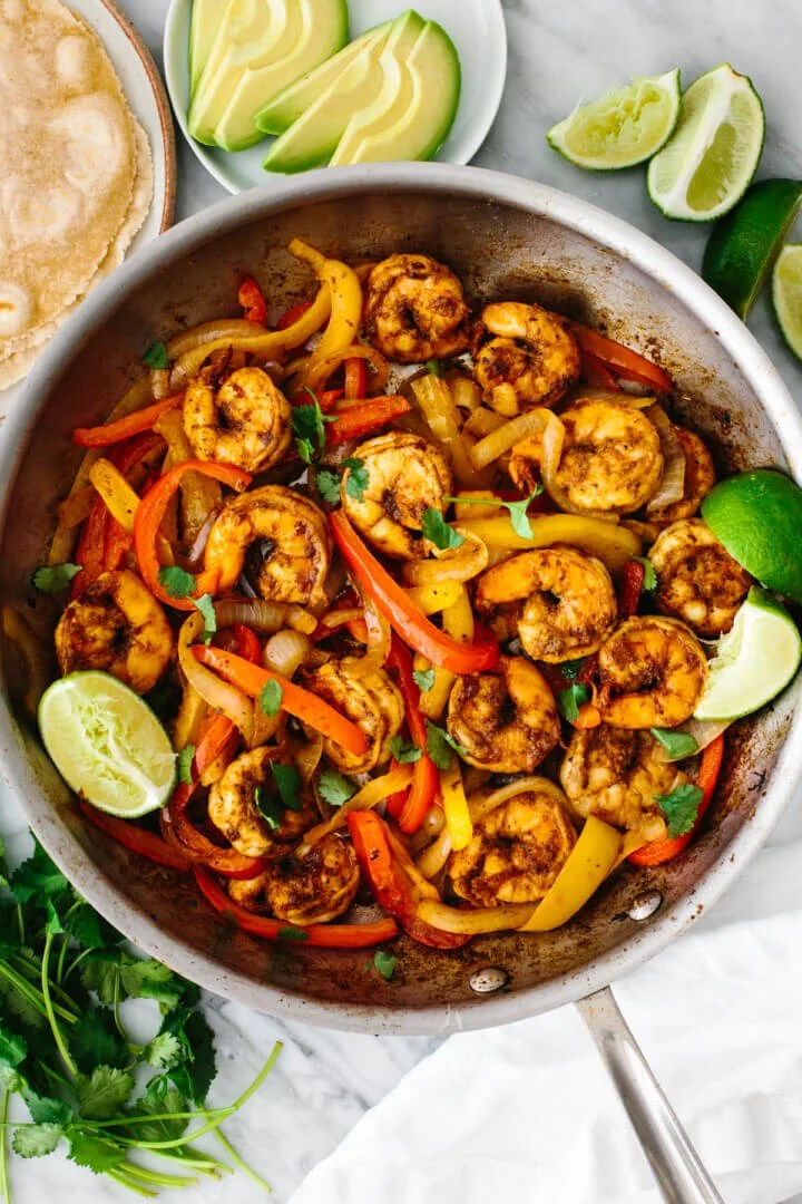 Shrimp fajitas in a pan with avocado slices and lime wedges.