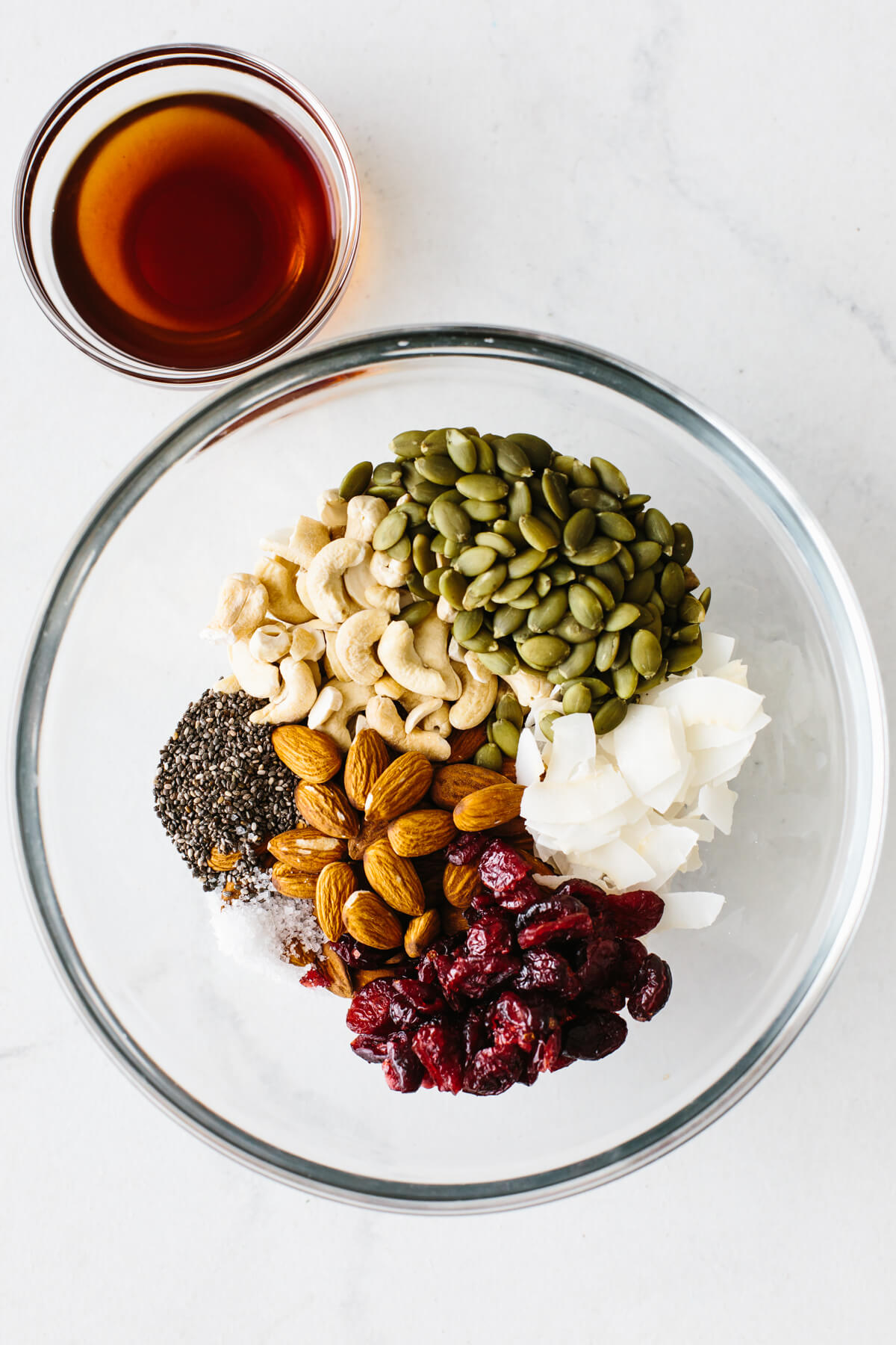 Trail mix granola bar ingredients in a mixing bowl, next to a smaller bowl of maple syrup.